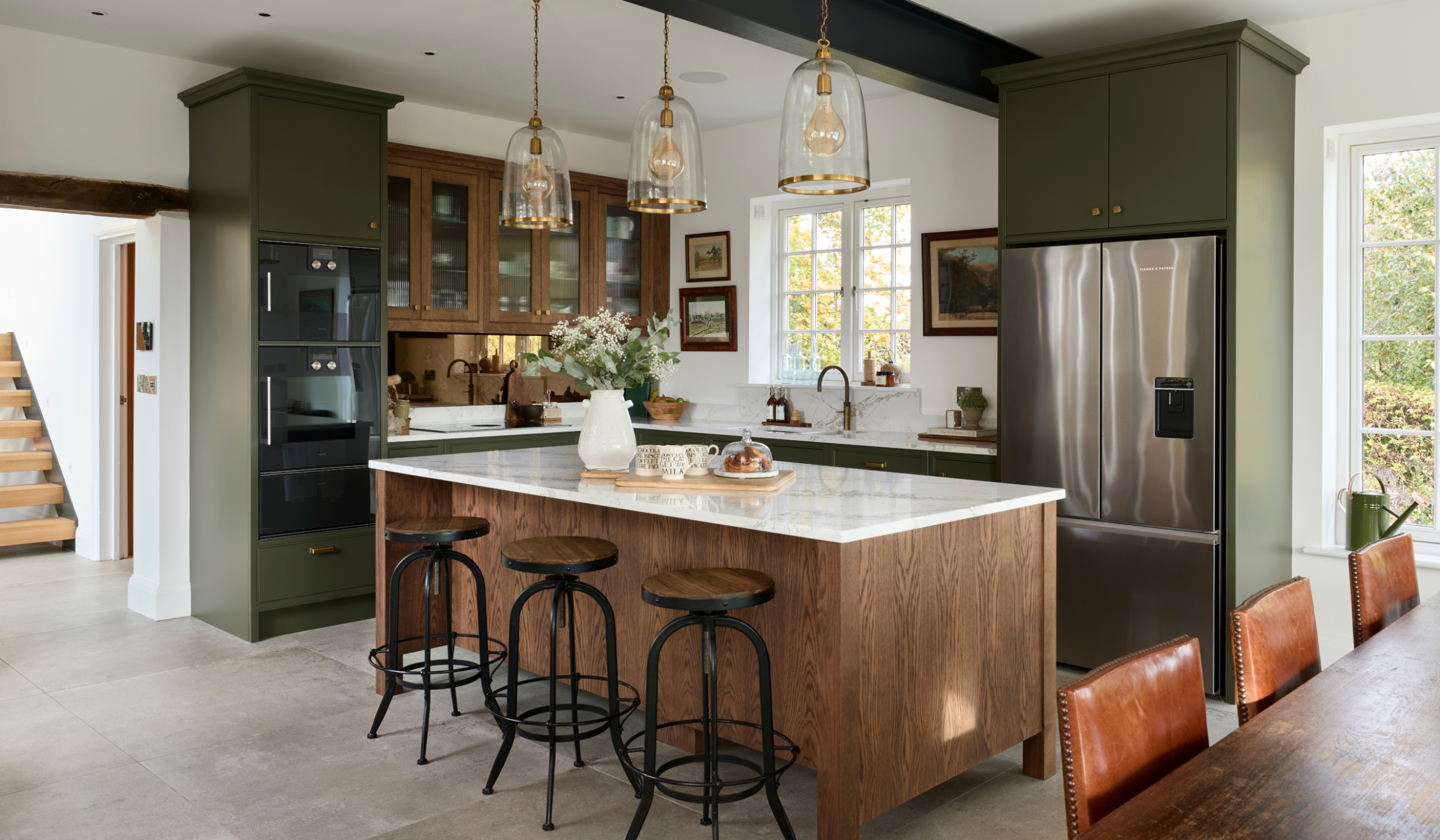 A traditional shaker kitchen in Invisible Green. The view of the raised central island with Verde Peake marble top is offset by the open kitchen doorway and the dining table with three chairs under it. On the marble top, a chopping board with three mugs and pain au chocolats stand next to a cream ceramic vase. To the left side of the central island, bespoke cabinetry stands from floor-to-ceiling and encloses premium Gaggenau appliances. Leading around from there, the Invisible Green theme continues with the cabinetry below the white worktop where a built-in hob catches the on eye on the dog-leg around to the right. Contrasting the Invisible Green, the darker wood finish of the cabinetry above the hob appears to oversee the patinated Quooker Fusion tap set into the worktop along the back wall. The worktop is finished off with a Fisher & Paykel fridge freezer with cabinetry surrounding it.