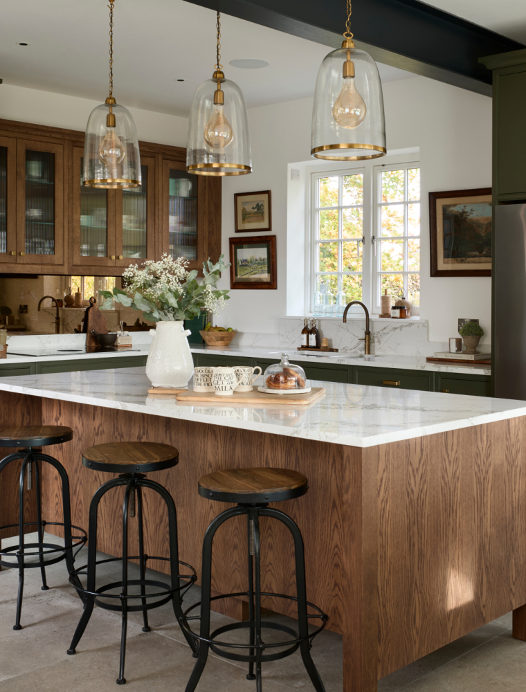 A traditional shaker kitchen in Invisible Green. A raised central island with Verde Peake marble top overshadows three black-legged stools with wooden seats. On the marble top, a chopping board with three mugs and pain au chocolats stand next to a cream ceramic vase. To the left side of the central island, bespoke cabinetry stands from floor-to-ceiling and encloses premium Gaggenau appliances. Leading around from there, the Invisible Green theme continues with the cabinetry below the white worktop where a built-in hob catches the on eye on the dog-leg around to the right. Contrasting the Invisible Green, the darker wood finish of the cabinetry above the hob appears to oversee the patinated Quooker Fusion tap set into the worktop along the back wall. The worktop is finished off with a Fisher & Paykel fridge freezer with cabinetry surrounding it.