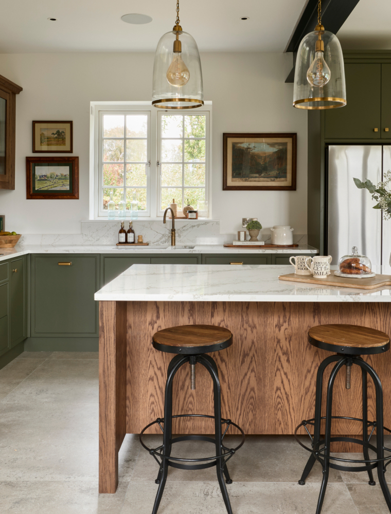 A traditional shaker kitchen in Invisible Green. A raised central island with Verde Peake marble top overshadows three black-legged stools with wooden seats. On the marble top, a chopping board with three mugs and pain au chocolats stand next to a cream ceramic vase. To the left side of the central island, bespoke cabinetry stands from floor-to-ceiling and encloses premium Gaggenau appliances. Leading around from there, the Invisible Green theme continues with the cabinetry below the white worktop where a built-in hob catches the on eye on the dog-leg around to the right. Contrasting the Invisible Green, the darker wood finish of the cabinetry above the hob appears to oversee the patinated Quooker Fusion tap set into the worktop along the back wall. The worktop is finished off with a Fisher & Paykel fridge freezer with cabinetry surrounding it.