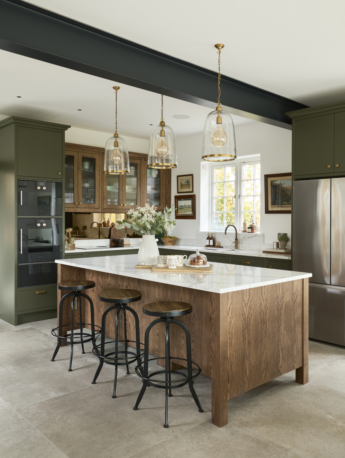 A traditional shaker kitchen in Invisible Green. A raised central island with Verde Peake marble top overshadows three black-legged stools with wooden seats. On the marble top, a chopping board with three mugs and pain au chocolats stand next to a cream ceramic vase. To the left side of the central island, bespoke cabinetry stands from floor-to-ceiling and encloses premium Gaggenau appliances. Leading around from there, the Invisible Green theme continues with the cabinetry below the white worktop where a built-in hob catches the on eye on the dog-leg around to the right. Contrasting the Invisible Green, the darker wood finish of the cabinetry above the hob appears to oversee the patinated Quooker Fusion tap set into the worktop along the back wall. The worktop is finished off with a Fisher & Paykel fridge freezer with cabinetry surrounding it.