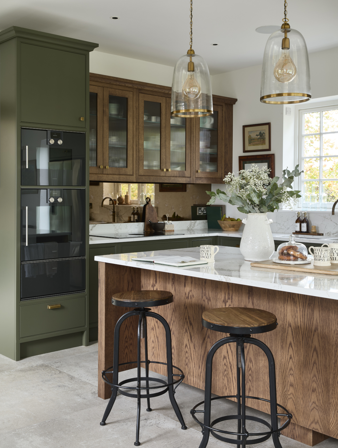 A traditional shaker kitchen in Invisible Green. A raised central island with Verde Peake marble top overshadows two black-legged stools with wooden seats. On the marble top, a chopping board with two mugs and pain au chocolats stand next to a cream ceramic vase and before an open book with mug next to it. To the left side of the central island, bespoke cabinetry stands from floor-to-ceiling and encloses premium Gaggenau appliances. Leading around from there, the Invisible Green theme continues with the cabinetry below the white worktop where a built-in hob catches the on eye on the dog-leg around to the right. Contrasting the Invisible Green, the darker wood finish of the cabinetry above the hob appears to oversee the patinated Quooker Fusion tap set into the worktop along the back wall.   