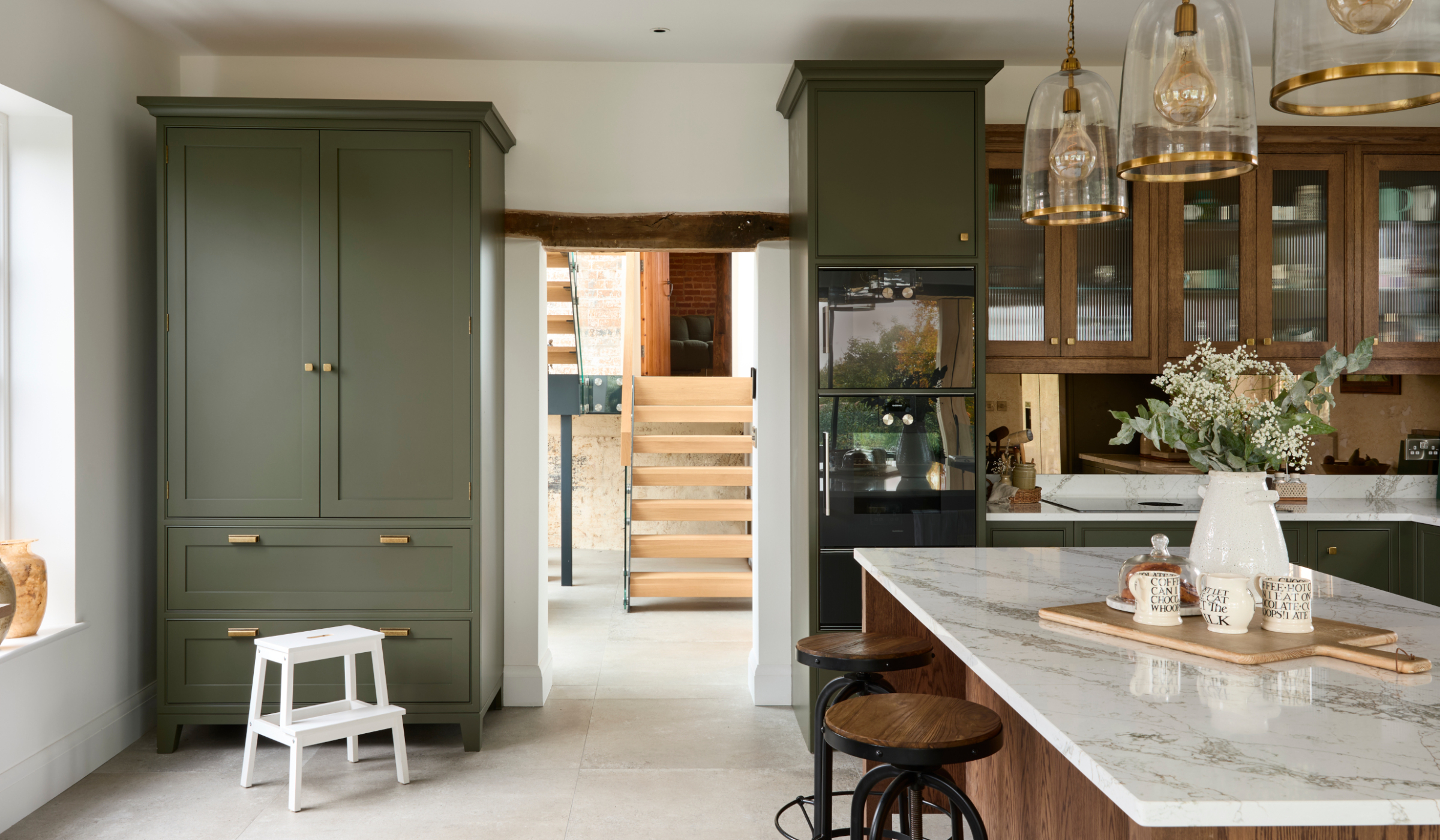 A traditional shaker kitchen in Invisible Green. A front on view of the open doorway out of the kitchen. On its left, a raised pantry with Antique Brass handles from Hendel & Hendel’s Otto range stands and on its right, tall bespoke cabinetry with premium Gaggenau appliances begins the cabinetry around the right fog leg of the room. The raised central island with Verde Peake marble top overshadows two black-legged stools with wooden seats and stands in line with the tall cabinet. 