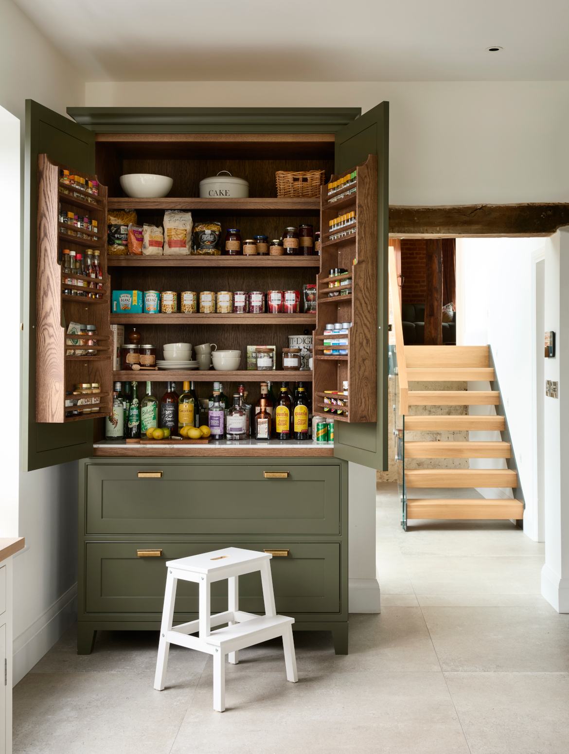 A traditional shaker kitchen in Invisible Green. A front on view of the open doorway out of the kitchen. On its left, a raised pantry with Antique Brass handles from Hendel & Hendel’s Otto range has its doors open revealing what’s inside. Inside, there are four shelves of savoury items such as pasta, beans and cooking oils. Two well-stocked spice rack adorns the backs of the cupboard doors. 