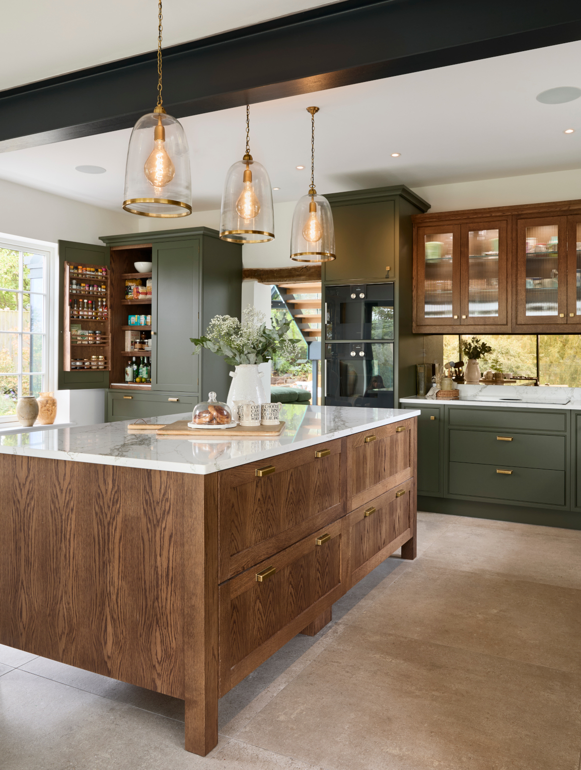 A traditional shaker kitchen in Invisible Green. The raised central island with Verde Peake marble top is overseen by tall bespoke cabinetry housing premium Gaggenau appliances. On the left side of the kitchen doorway, a raised pantry with Antique Brass handles from Hendel & Hendel’s Otto range has an open door revealing well-stocked shelves and spice cabinet within. 