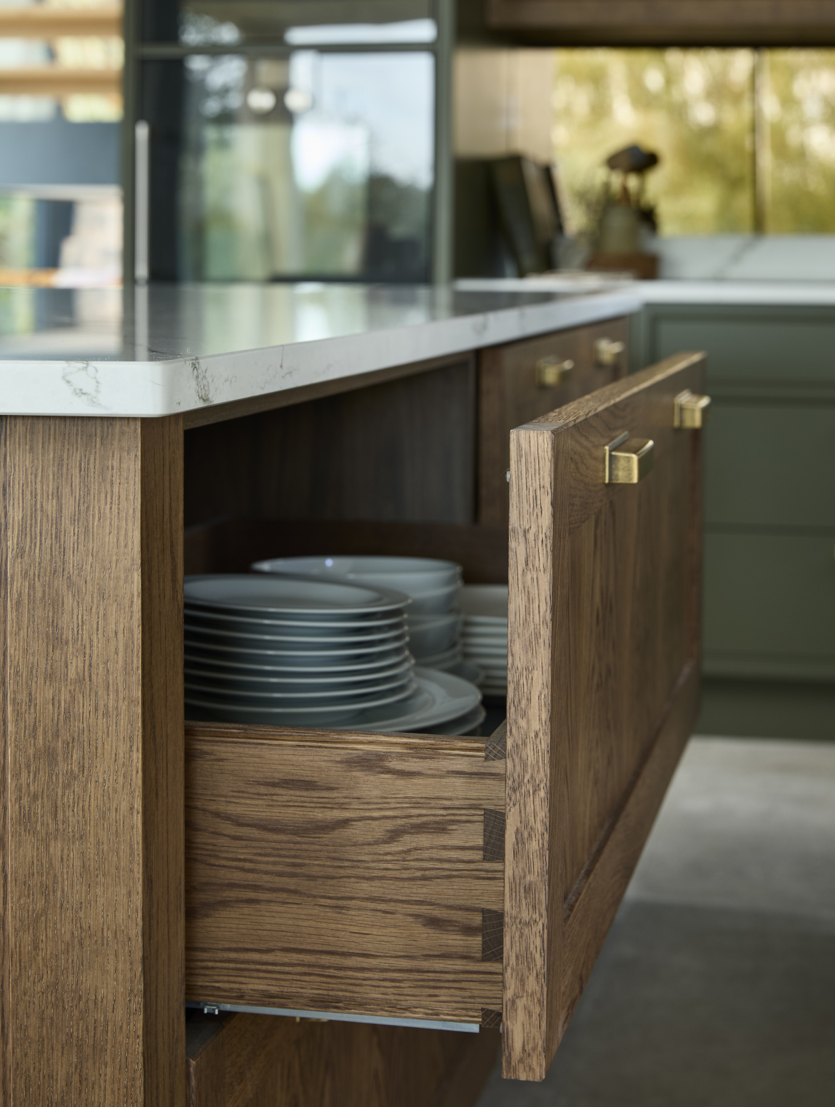 A traditional shaker kitchen in Invisible Green. A close in view of an open drawer from the raised central island. The Verde Peake marble top contrasts with the darker wood finish of the drawer itself. The wood finish compliments the Antique Brass handles from Hendel & Hendel’s Otto range on the drawer. Inside the drawer, a selection of different plates can be seen. 