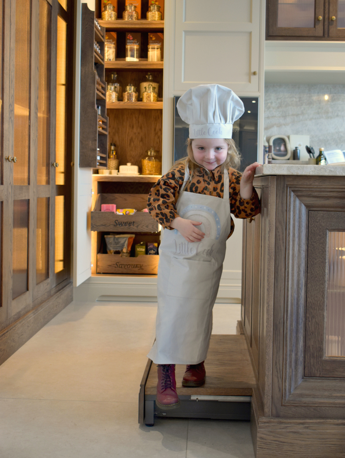 A young girl dressed in chef's attire stands on a raised plinth pulled out from kitchen island.