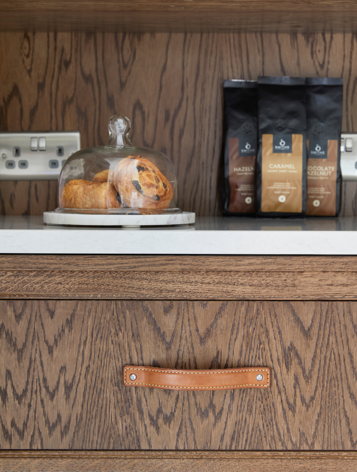 A traditional shaker kitchen in French Grey. The bottom shelf of the bespoke floor-to-ceiling pantry has selection of coffees and a dish of breakfast pastries. The well-joined drawer below contrasts with the white worktop.